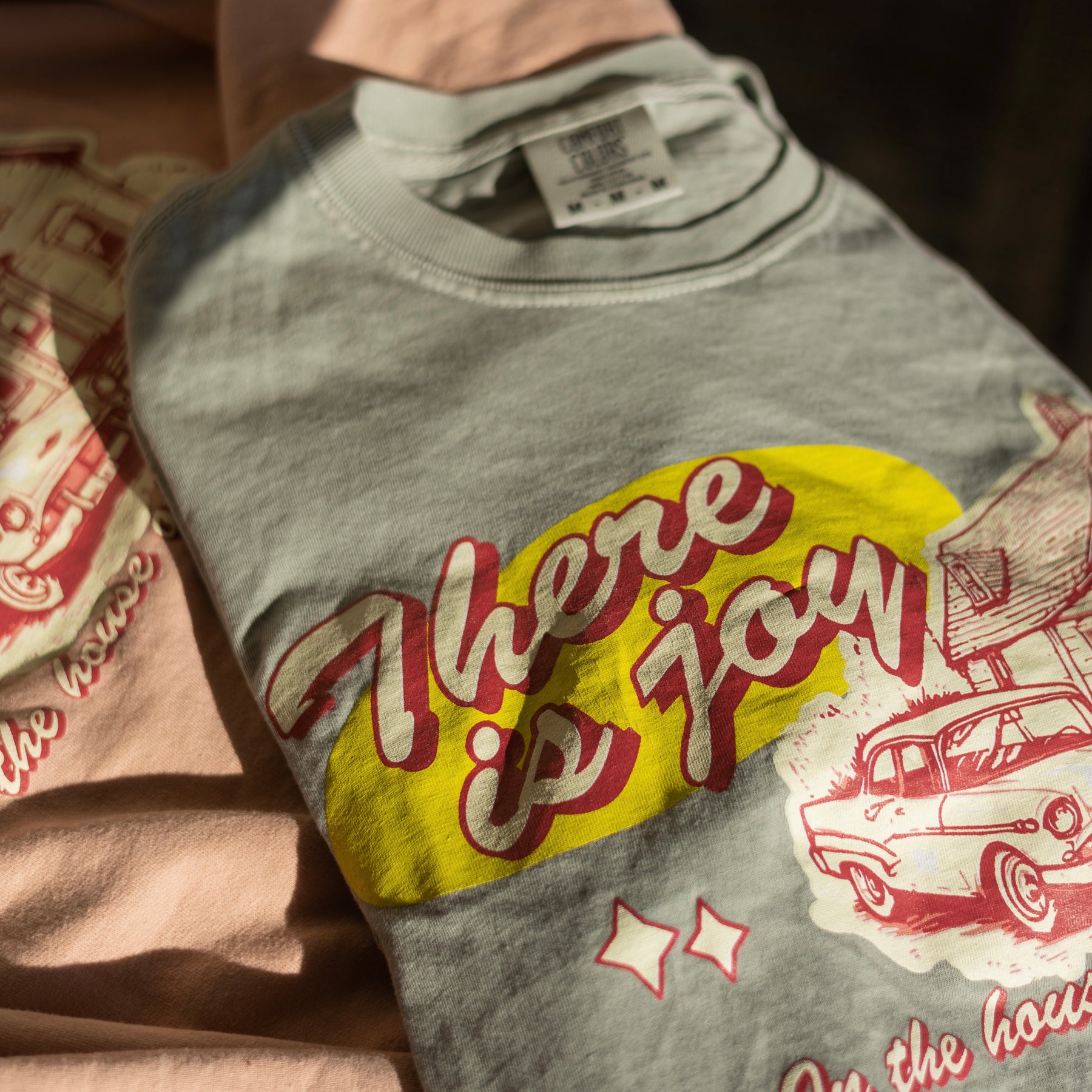Neutral sage green t-shirt with yellow and red graphic design on a wooden surface