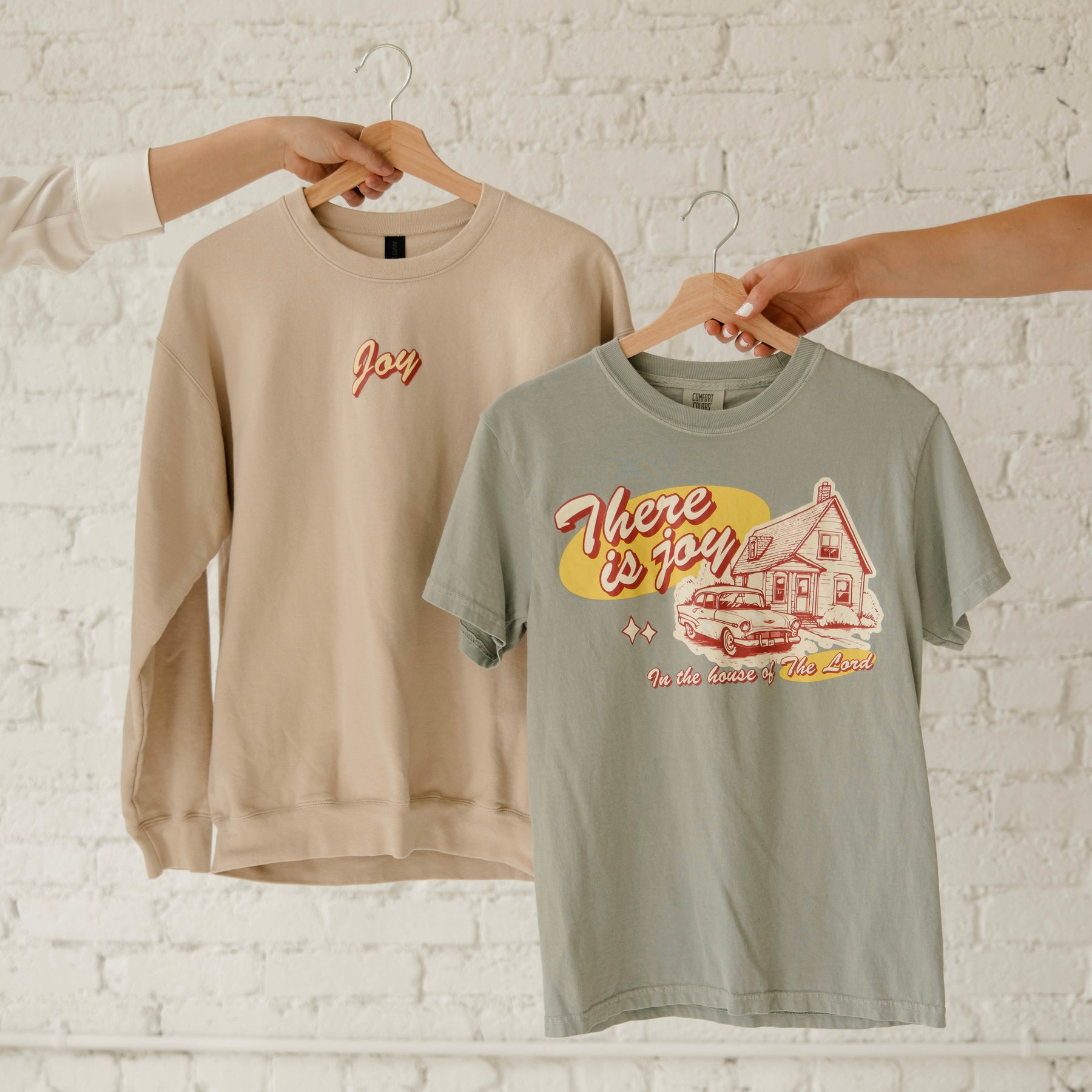 Beige Christian "joy" sweatshirt and green t-shirt with graphic designs on a white brick wall background