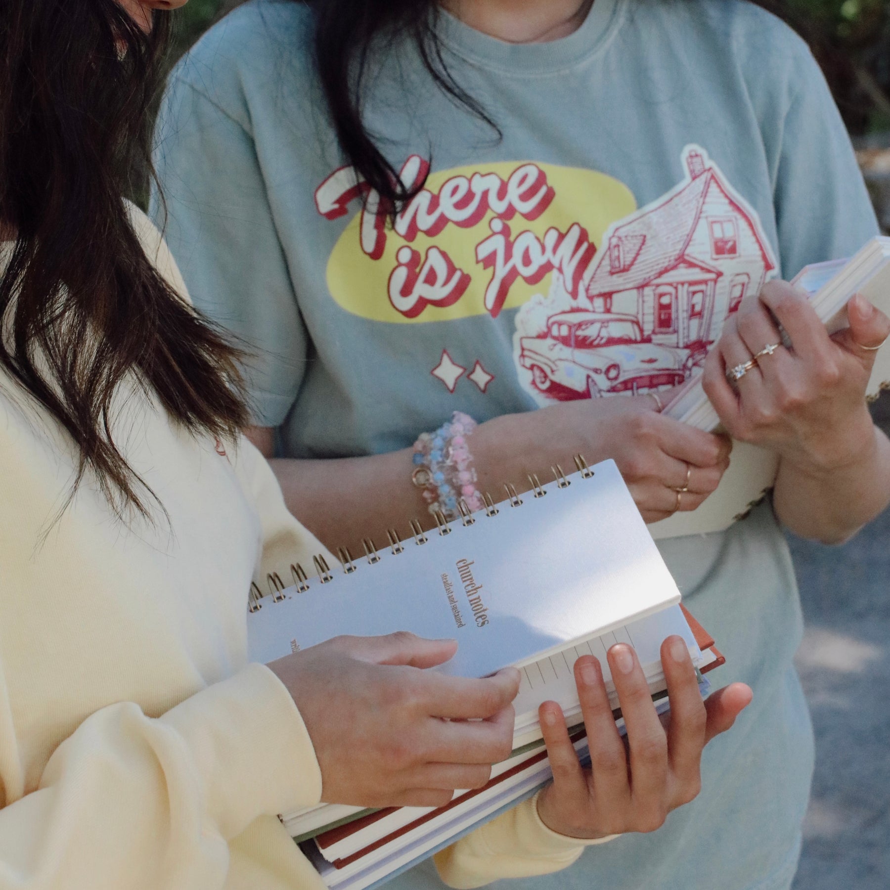 Two people holding church notebooks with a person wearing a shirt with text and vintage graphics, saying there is joy in the house of the Lord.
