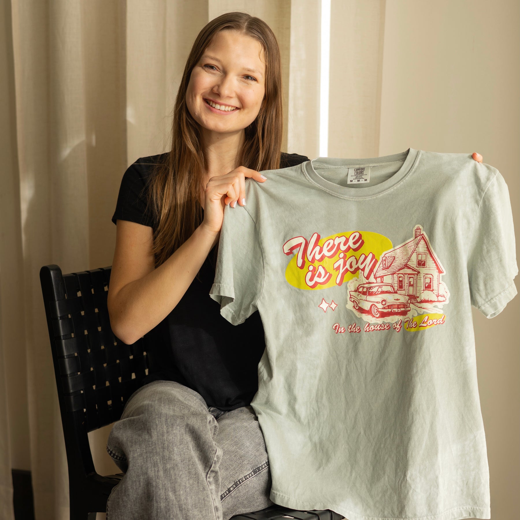 Woman holding a t-shirt with a vintage graphic design of a house and car, and text saying there is Joy in the house of the Lord