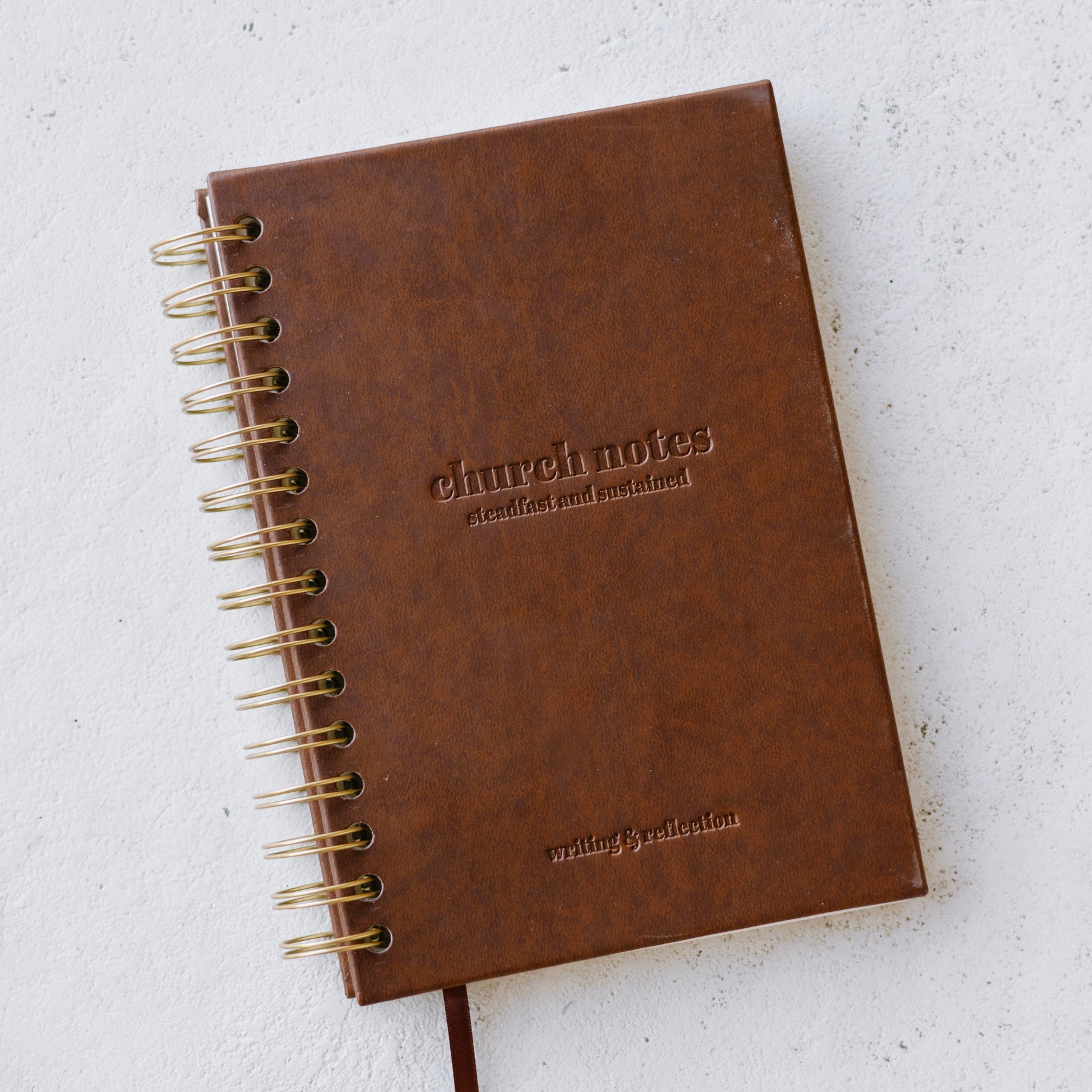 Brown faux leather notebook with 'church notes' 'steadfast and sustained' 'writing & reflection' embossed on it, on a light gray background