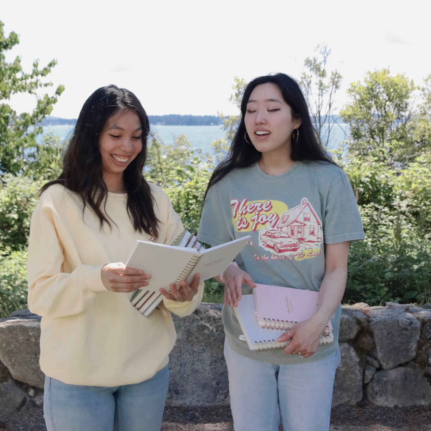 Two women standing outdoors by a lake, holding spiral linen church notebooks, and wearing a Christian merch tee saying there is joy in the house of the Lord