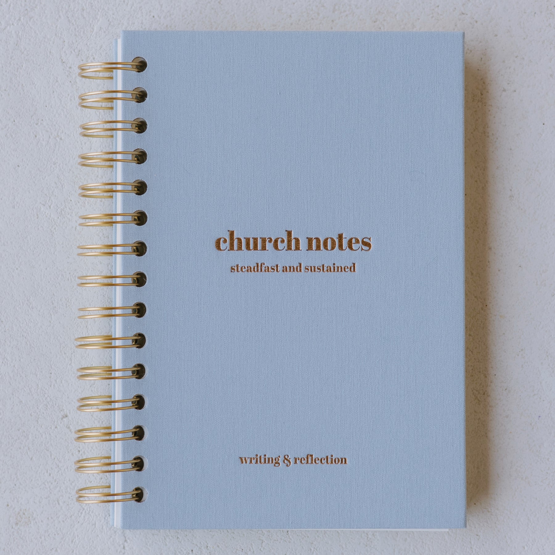 Light blue notebook with gold text on a light gray background, with text reading 'church notes' 'steadfast and sustained' 'writing & reflection' 